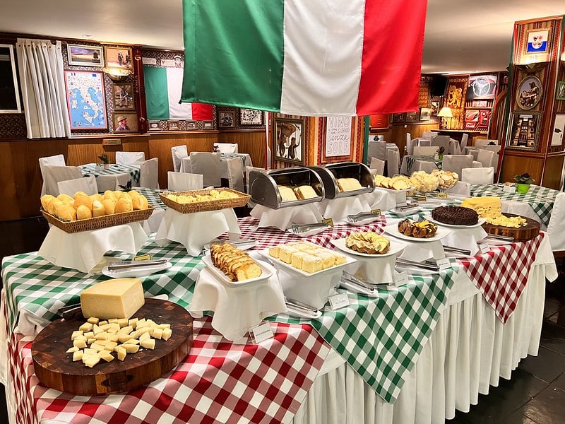 Buffet de café da manhã no Hotel Bella Italia, com uma grande variedade de pães, queijos e doces dispostos em uma mesa decorada com toalhas de mesa xadrez em verde, branco e vermelho, representando a cultura italiana. A bandeira da Itália está pendurada acima da mesa, criando uma atmosfera acolhedora e temática para os hóspedes apreciarem as delícias matinais.