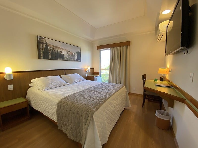 Quarto de hotel confortável e acolhedor em Foz do Iguaçu, com cama de casal, decoração elegante, além de uma iluminação suave. O ambiente conta com ar-condicionado, mesa de trabalho e uma vista agradável da janela, ideal para quem busca descanso após explorar as atrações turísticas da região. Este hotel oferece as melhores tarifas em Foz do Iguaçu, proporcionando conforto e praticidade para seus hóspedes em uma localização estratégica.
