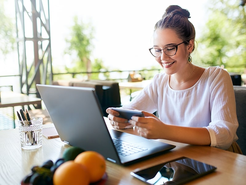 Mulher sorrindo enquanto utiliza o Wi-Fi em um hotel com ambiente ao ar livre em Foz do Iguaçu. Com o laptop e celular em mãos, ela aproveita o Wi-Fi gratuito oferecido pelo hotel, ideal para quem busca trabalhar ou se conectar em um hotel com Wi-Fi em Foz, sem abrir mão do conforto e da natureza ao redor.