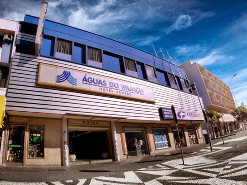 Fachada do hotel Águas do Iguaçu, com estrutura moderna e linhas horizontais em tons de branco e azul. O nome do hotel está destacado em uma placa centralizada na parte superior da entrada. O prédio possui janelas retangulares escuras na parte superior, e a entrada principal é ampla, com pilares laterais. Ao lado, uma farmácia é visível, e a calçada em frente ao hotel apresenta um padrão geométrico. O céu azul e claro complementa o cenário urbano.