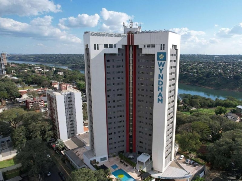 Vista aérea do hotel Wyndham Golden Foz Suites, em um edifício moderno e alto, com fachada branca e cinza, situado em uma localização privilegiada próxima ao rio. O hotel oferece acomodações de luxo, piscina externa e vistas panorâmicas da cidade e da natureza ao redor. Ideal para viajantes que procuram conforto, serviços de qualidade e fácil acesso às atrações de Foz do Iguaçu.