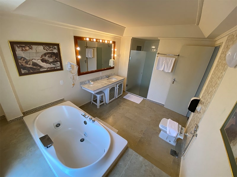 Banheiro espaçoso e luxuoso de um hotel em Foz do Iguaçu, equipado com uma banheira de hidromassagem, pia dupla e um espelho grande iluminado. A decoração elegante proporciona um ambiente relaxante, perfeito para momentos de conforto e bem-estar. Este hotel, com as melhores tarifas na cidade, oferece comodidades de alto padrão para hóspedes que buscam uma experiência sofisticada e agradável durante sua estadia.