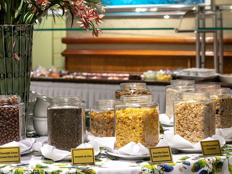 Mesa de café da manhã de um hotel, com uma grande variedade de cereais, incluindo sementes de chia e sucrilhos, organizados em potes de vidro. Este hotel, conhecido por oferecer as melhores tarifas em Foz do Iguaçu, também se destaca pela qualidade e diversidade de seu buffet matinal, ideal para quem procura começar o dia com uma refeição saudável e saborosa antes de explorar a cidade e suas atrações.