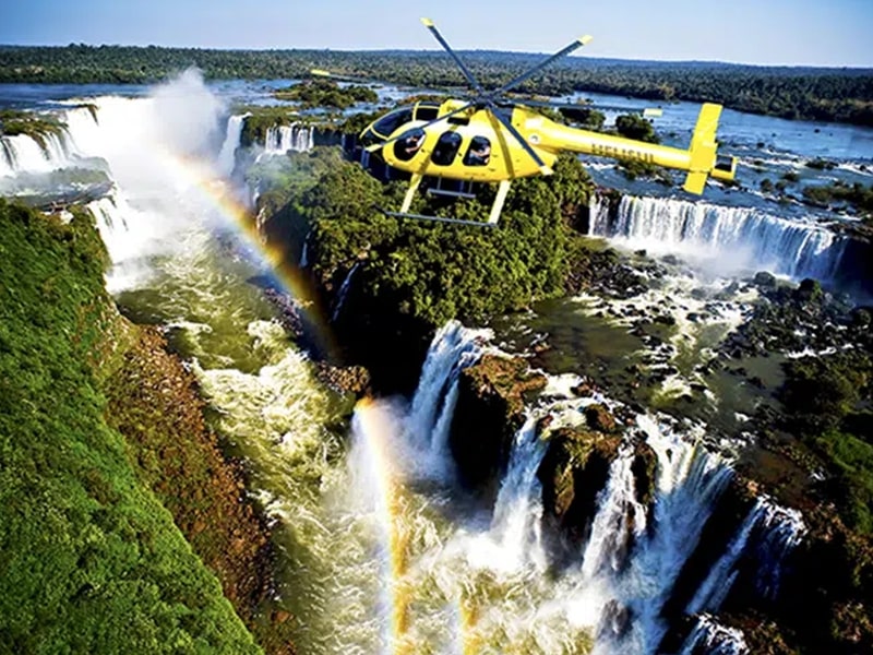 Imagem de um helicóptero amarelo sobrevoando as Cataratas do Iguaçu, oferecendo uma vista panorâmica deslumbrante. Um arco-íris se forma sobre as quedas d'água, adicionando um toque mágico à paisagem exuberante, onde a força das águas e a vegetação ao redor criam uma experiência visual impressionante.