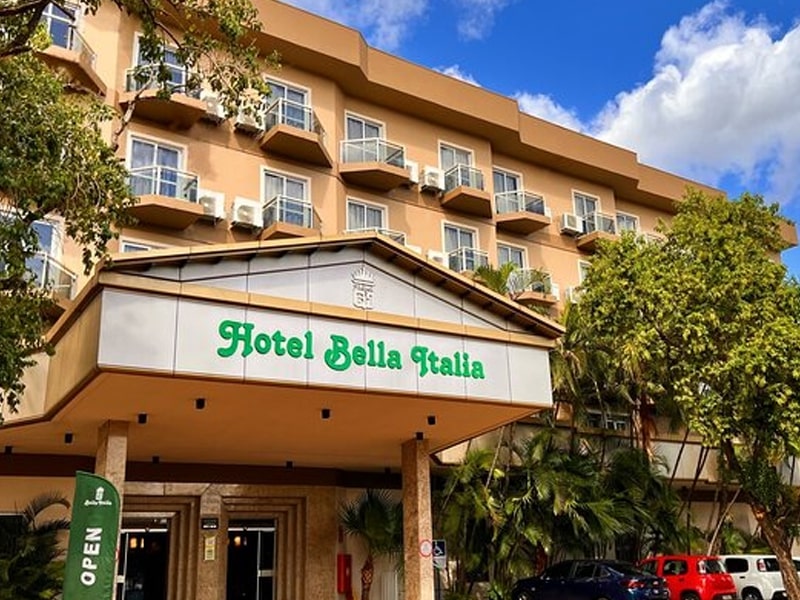 Fachada do hotel Bella Italia com estrutura de três andares e varandas em cada quarto, com plantas decorativas ao redor da entrada. A entrada principal possui um letreiro com o nome do hotel em letras verdes, em uma área coberta com colunas e detalhes arquitetônicos em tons neutros. Árvores e vegetação cercam a área, contribuindo para uma atmosfera acolhedora e agradável. O céu azul e algumas nuvens completam o cenário, transmitindo uma sensação de tranquilidade e conforto.