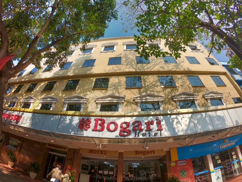 Fachada do Bogari Hotel, com uma estrutura em tons claros e janelas retangulares distribuídas de maneira simétrica. O letreiro com o nome do hotel está em destaque na entrada, com letras vermelhas e um símbolo floral. Árvores com folhagem densa cercam o edifício, projetando sombras sobre a fachada e criando um ambiente agradável e convidativo. A calçada em frente ao hotel é movimentada, e lojas próximas, como uma farmácia, são visíveis na lateral.