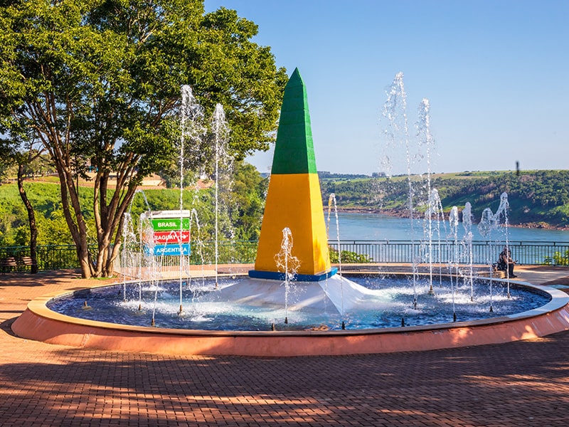 Marco das Três Fronteiras em Foz do Iguaçu, com um obelisco colorido em verde, amarelo e azul, representando o Brasil. Ao redor do obelisco, uma fonte com jatos de água oferece um visual agradável e refrescante, cercada por árvores e vegetação nativa. Ao fundo, a vista panorâmica do rio Paraná separa o Brasil da Argentina e do Paraguai, simbolizando a união das três nações em um dos pontos turísticos mais emblemáticos da região.