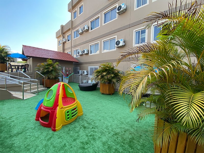 Área externa infantil de um hotel em Foz do Iguaçu, com brinquedos coloridos e um espaço seguro e confortável para as crianças se divertirem. O ambiente é cercado por plantas, criando uma atmosfera agradável e tranquila. Este hotel, com as melhores tarifas da cidade, oferece opções de lazer para toda a família, garantindo que todos, inclusive os pequenos, tenham uma estadia prazerosa enquanto desfrutam das atrações da região.