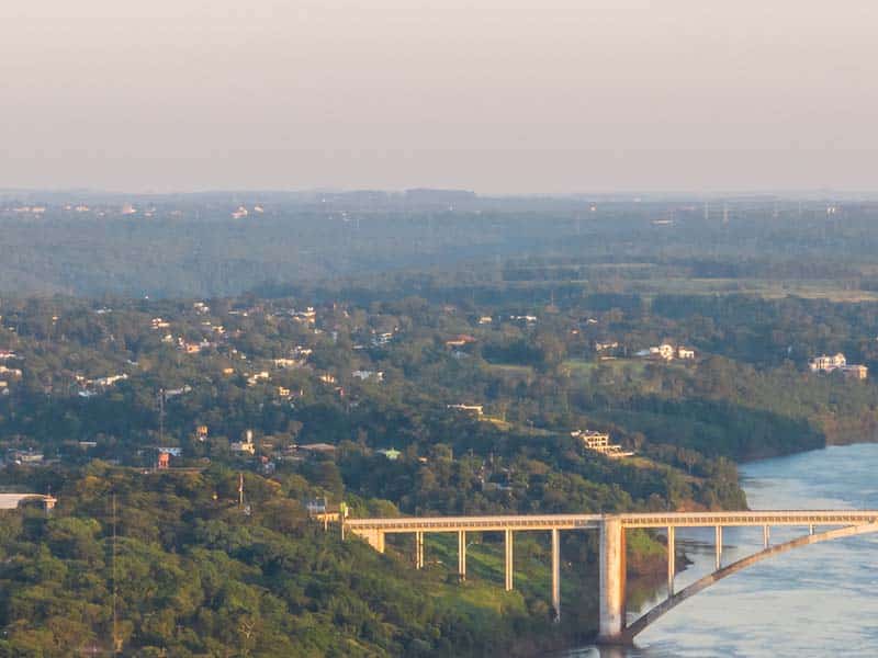 Vista panorâmica da Ponte da Amizade ao entardecer, com uma ampla extensão de vegetação ao fundo, onde pequenas construções e casas estão distribuídas entre as áreas verdes. A ponte, uma estrutura em arco sobre o rio, conecta duas margens densamente arborizadas. A luz suave do fim do dia ilumina a paisagem, destacando a ponte e o contraste entre o rio e a vegetação ao redor, criando uma sensação de serenidade e imensidão.